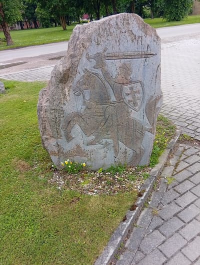 A stone monument with a carved image of a knight on horseback is situated in front of a paved walkway and a grassy area with trees in the background. The knight is depicted wearing armor and holding a sword, with a shield bearing a cross symbol. Small yellow and white flowers are growing at the base of the monument. The monument appears to be located in the city of Micaičiai, Lithuania.