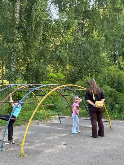 The image depicts a playground scene in Sarov, Russia, under a canopy of lush green trees, possibly birch trees due to their distinctive white and black bark visible in the upper left portion of the image. In the foreground, a metal jungle gym with colorful blue, red, and yellow bars stands on an asphalt surface. Three individuals are present: a young boy in a turquoise t-shirt and black pants is hanging from the jungle gym; a young girl in a pink t-shirt and light blue jeans is standing and looking towards the viewer, with her hand raised to her head; and a woman with long brown hair, wearing a black t-shirt and dark brown flared pants, stands facing away from the viewer, with a yellow crossbody bag slung over her shoulder. The environment appears to be a park or a residential area with abundant greenery and dappled sunlight.