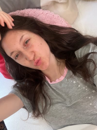 The image shows a close-up, overhead shot of a young woman lying down. She has long, dark brown hair that is spread out around her. Her head is tilted, and she is making a kissing face towards the camera. She is wearing a gray t-shirt with white stars on it, and a pink trim around the neckline. Her skin appears to have freckles and some minor blemishes. She has her left hand resting on her head, with her fingers slightly spread. The background consists of pillows and bedding. The lighting suggests an indoor environment, possibly a bedroom. There is no indication of the location being in the city of Popintsi, Bulgaria.