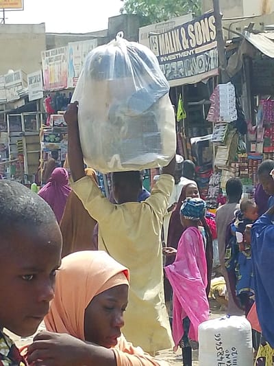The image depicts a crowded outdoor market scene in Fagge D1, Nigeria, under bright daylight. In the mid-ground, a person, viewed from behind and wearing a light-colored long-sleeved garment, carries a large, clear plastic bag filled with various items on their head, stabilizing it with both hands raised. To the left foreground, the left profile of a young male with short dark hair, wearing a patterned shirt, is visible, looking towards the right. To his right, a female figure in an orange headscarf is partially visible, looking downwards.
Numerous individuals populate the mid-ground and background, indicating a bustling environment. Among them, a person in a purple head covering is seen to the left of the central figure, and a woman in a bright pink headscarf and matching garment is positioned to the right. Further right, a person in blue clothing holds a baby wrapped in patterned fabric. In the background, multiple market stalls are visible, featuring goods displayed on shelves. A prominent sign above one stall reads "MALIK & SONS INVESTMENT" in white lettering on a dark background. Another white sack with the black inscription "9785466 KANO 111120" rests in the lower right foreground. The ground appears unpaved and dusty.