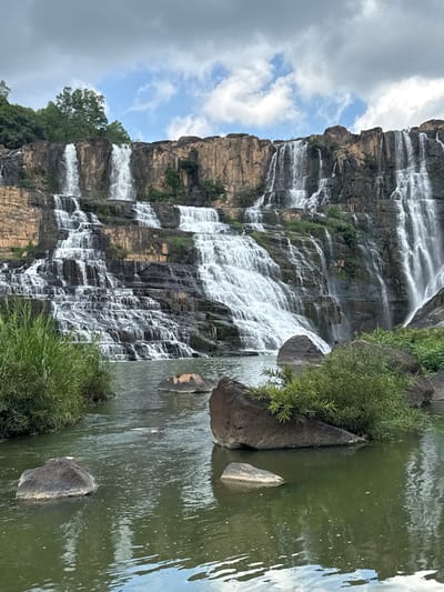 The image shows the Pongour Waterfall in Tân Phú, Vietnam. The waterfall cascades down a series of large, tiered rock formations. The water flows in multiple streams, creating a white, frothy appearance as it tumbles over the rocks. The surrounding environment is lush and green, with tall grass and trees growing on the banks of the river at the base of the waterfall. Several large, dark rocks are scattered in the water, some partially submerged. The sky above is partly cloudy, with a mix of blue sky and grey clouds. The overall scene depicts a powerful and natural landscape.