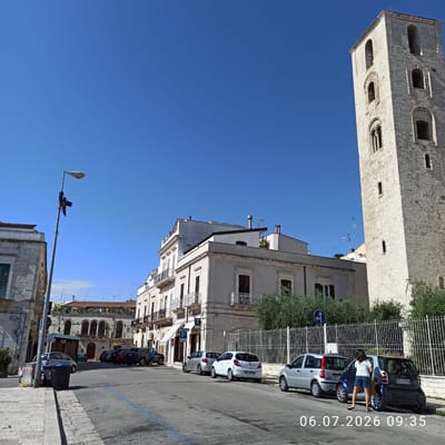The image captures a sunlit street scene in Ruvo di Puglia, Italy, with a prominent bell tower on the right side. The tower, made of light-colored stone, features multiple arched windows and appears to be part of a historic building, likely a church. The street is lined with parked cars, including a silver compact car, a white hatchback, a dark-colored SUV, and another silver compact car closer to the foreground. Two women are standing near the parked cars; one is facing away from the camera, wearing a white t-shirt and blue shorts, while the other is partially visible, wearing a light-colored top and dark bottoms. A lamppost with lights and what appears to be a camera is visible on the left side of the street. The buildings lining the street are a mix of architectural styles, with some featuring balconies and awnings. The sky is a clear, bright blue, indicating good weather conditions. The timestamp "06.07.2026 09:35" is superimposed on the lower right portion of the image. In the foreground, there are trash receptacles and a blue painted line on the asphalt, possibly indicating a parking zone.