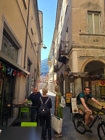 In Bellinzona, Switzerland, on a sunny day, a narrow street is lined with buildings adorned with flags. A man with a shaved head stands near a table with a glass of rosé and an appetizer, talking to a woman with blonde hair and a backpack. To the right, a man with glasses rides a bicycle. The street is paved, and shops line the buildings, some with signs in Italian. The atmosphere suggests a leisurely afternoon in a European town.