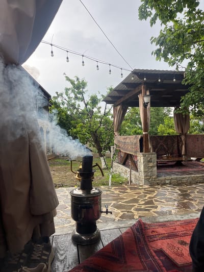 The scene is an outdoor area in what appears to be a leisure setting, possibly a backyard or an outdoor cafe in Miatli, Russia. The central object is a samovar, a metal container traditionally used to heat water for tea. White smoke emanates from it. The samovar sits on a wooden table. In the background, there's a covered structure with stone pillars and a wooden roof. The structure is decorated with rugs or tapestries. The ground around the structure is paved with irregular stone slabs. The sky is overcast. A string of lights hangs overhead on the left side of the frame. In the bottom right of the frame are a pair of shoes. A tree and other greenery are present in the middle ground. A tan tent or canopy wall is in the foreground.