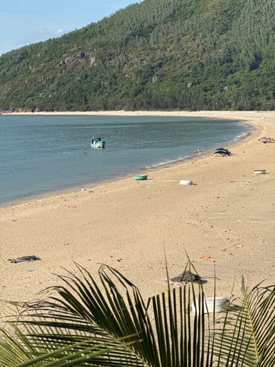 A wide shot captures a serene coastal scene at the city of Song Cau, Vietnam, featuring a sandy beach meeting calm, turquoise waters under a clear blue sky. A lush green, densely forested mountain range dominates the background, its slopes dotted with visible rocks and trees. In the foreground, the sandy beach is littered with various objects, including several small, upturned boats, some with white interiors, and a larger, dark, covered object near the water's edge, possibly a boat or shelter. A few scattered pieces of debris, including a piece of orange plastic and some dark netting, are visible on the sand. A single, small, light-colored fishing boat is anchored in the shallow water, with what appear to be buoys or floats trailing behind it. In the far distance, a few small figures and a small cart with yellow elements are visible on the beach. The lower portion of the image is partially obscured by the out-of-focus fronds of a palm tree.