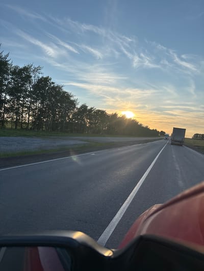 This is a photograph taken from a vehicle, likely a car, looking out at a highway at sunset. The perspective is from the passenger side, with the side mirror and a portion of the car's body visible in the foreground on the right. The road stretches into the distance, marked by a white line on the right lane. Several trucks are visible on the highway, with one in the middle distance and others further ahead.
To the left of the road, a dense line of trees borders the highway, with the setting sun peeking through them, casting a warm, golden light. The sky above is a vibrant blue, streaked with wispy white clouds that are illuminated by the setting sun. The overall impression is one of a peaceful journey during the golden hour, with the natural beauty of the sky and trees contrasting with the man-made road and vehicles. The image evokes a sense of travel and contemplation. The location cues suggest that this scene is taking place in Таймурзино, Russia.