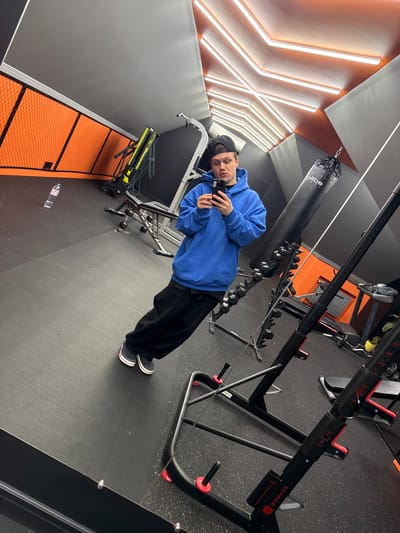A person wearing a blue hooded sweatshirt, black pants, and a baseball cap is standing in what appears to be a gym. The person is holding a cell phone and appears to be taking a picture or video. A punching bag and weightlifting equipment are visible in the background. The location is Шемякино, Russia.