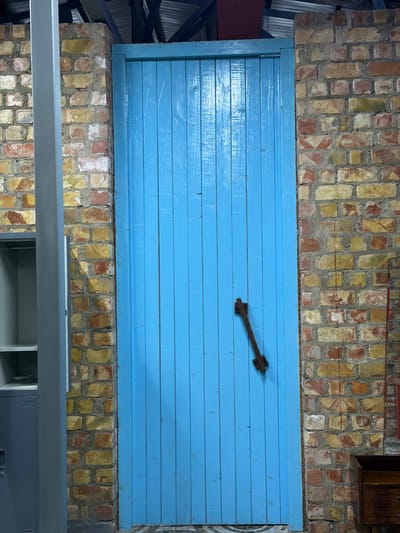 A bright blue wooden door with vertical planks is set into a brick wall. The brickwork is rustic, with a mix of red, yellow, and white bricks. A dark wooden latch mechanism is attached to the door at a diagonal. To the left of the door, a grey metal pillar stands adjacent to the brick wall, and a portion of a grey metal cabinet with open shelves is visible. The ceiling above shows exposed metal beams and what appears to be insulation or roofing material. The floor is tiled. The scene is located in the city of Shabdan, Kyrgyzstan.