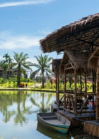 A wide-angle vertical shot captures a serene scene at a resort in Bintang Meriah, Indonesia, under a bright blue sky with wispy clouds. The foreground is dominated by a calm body of water reflecting the lush greenery and towering palm trees that line its edge. A small, weathered rowboat, painted with blue and green accents, rests gently on the water's surface, tethered to a wooden dock.

Adjacent to the water, a series of open-air pavilions with thatched roofs and bamboo structures offer shaded seating. These structures are built on a wooden platform that extends over the water. In the middle ground, a path winds through vibrant green foliage and decorative plants, with several palm trees scattered throughout.

Further in the background, a roller coaster structure with red and white tracks is visible, hinting at a recreational park or amusement area. Three individuals, dressed in red and dark clothing, are walking along the path near the water's edge, their reflections faintly visible on the calm surface. The overall impression is one of a tranquil, tropical getaway with elements of both natural beauty and recreational activities.