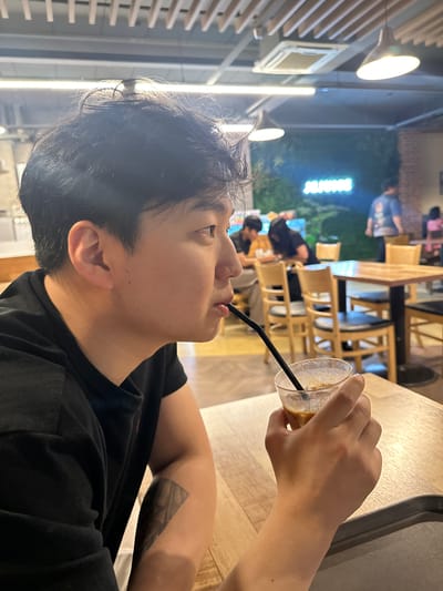 In a cafe, a young East Asian man with short, dark hair is seated at a wooden table, holding a clear glass with a brown beverage and a black straw to his lips. He is wearing a black t-shirt and has a tattoo on his left forearm. The cafe has wooden tables and chairs, with a neon sign that reads "JUHJUHSE" on a green wall in the background. Other patrons are visible in the background, some seated at tables. The lighting is a mix of overhead fluorescent lights and warm pendant lamps. The overall setting appears to be casual and relaxed. The location is the JUHJUHSE cafe in Dangjin-si, South Korea.