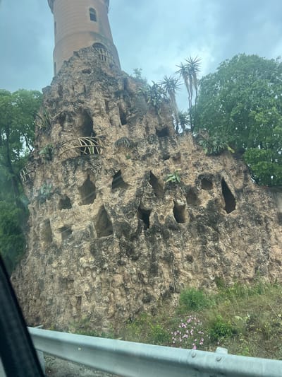 The image captures the architectural oddity of the Torre del Port in Cambrils, Spain. The structure features a tower constructed of what appears to be stacked stone, interspersed with rectangular hollowed-out openings resembling a dovecote. Cacti and green foliage grow from some of the openings. A section of what appears to be metal guardrail is visible in the foreground and side of the image, suggesting the vantage point is from an elevated position, likely a vehicle. Overcast sky dominates the top of the frame, with greenery at the top and bottom.