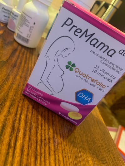 This is a close-up, angled photograph of a box of "PreMama" prenatal vitamins, likely taken indoors on a wooden surface. The box is predominantly pink and white and features a stylized illustration of a pregnant woman. Prominent text on the box includes "PreMama," "preconception, pregnancy & breast-feeding," "11 vitamins 10 minerals," and "Quatrefolic active folic acid - folate." A hexagonal blue badge displays "DHA," and the side of the box indicates "30 tablets 30 soft capsules Net weight: 57.75 g" and "food supplement." Two white bottles, one with a pink cap, are partially visible in the foreground to the left. The background is blurred but suggests an indoor setting with some shelving. The lighting appears to be artificial, casting a warm tone on the wooden surface. The scene is static, focusing on the product packaging. No specific time of day, weather, or emotional cues are discernible. The provided location context, Krushev Dol, Bulgaria, is not visually evident in the image itself.