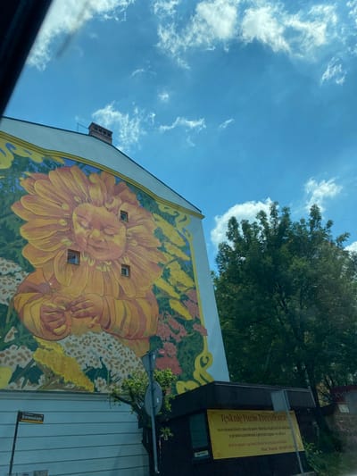 The image captures a vibrant mural on the side of a building in Bielsko-Biała, Poland. The mural depicts a stylized, childlike face within a large, anthropomorphic sunflower, surrounded by lush foliage and flowers. The artwork appears to be a cheerful and whimsical representation. Below the mural, a street sign indicates "ul. Władysława Broniewskiego". To the right, a small, dark-colored building with a yellow banner is visible. The banner features Polish text, which appears to be an advertisement or announcement, and a telephone number. A mature tree with dense green foliage stands beside the building. The sky above is a bright blue with scattered white clouds, suggesting a clear day. The overall scene is urban, with elements of art, nature, and street infrastructure.