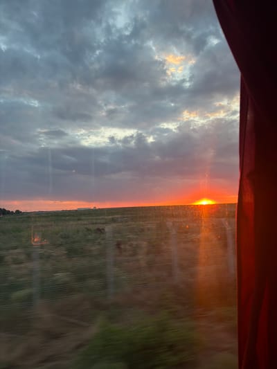 This image depicts a sunset over a rural landscape, likely in or around Sibioara, Romania. The sun, a brilliant orange orb, is low on the horizon, casting a warm glow across the scene. It is partially obscured by a horizontal line of dark clouds, with patches of lighter clouds above, tinged with pink and yellow. The sky transitions from a deep orange at the horizon to shades of purple and gray higher up.

In the foreground, a field of green and brown vegetation stretches towards the horizon. A series of thin, white horizontal lines, possibly wires or fences, are visible across the field, suggesting a man-made element within the natural landscape. The vegetation appears somewhat blurred, indicating movement, possibly from a moving vehicle.

On the far horizon, a few structures are visible, including what appears to be a white building or silo, and possibly distant trees or power lines. The right side of the image is dominated by a dark red curtain, which frames the view of the sunset. The curtain is opaque and slightly wrinkled, and it partially obscures the sky and the landscape. The overall impression is one of a peaceful, natural setting at dusk, viewed from a slightly obscured vantage point.