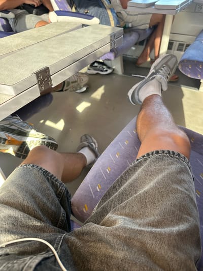The image depicts the interior of a train carriage, focusing on the legs and feet of passengers. In the foreground, a person is seated, wearing grey denim shorts and white socks with grey sneakers. Another person's legs and feet, wearing grey shorts and black and white sneakers, are visible slightly further back. Other passengers are partially visible in the background, seated and with their legs extended. The train carriage features grey folding tables with silver hinges, and the seats are upholstered in a blue fabric with yellow and white stripes. The flooring is a light grey. The overall environment suggests travel, possibly within a city or between locations. The specific location in the city of Salses-le-Château, France, is not discernible from the visual information present in the image.
