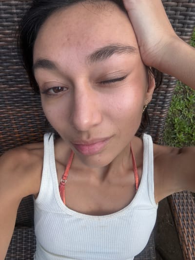 A forensic examination of the image reveals a female individual reclining outdoors in Hanalei, United States of America. The subject is depicted from a high-angle, close-up perspective, looking directly into the camera with a wink; her right eye is open, while her left eye is closed. Her right hand is placed on the upper right side of her head, partially obscuring her hair. She possesses dark hair, a medium skin tone, and prominent eyebrows. Her lips are slightly parted.
The subject is wearing a white ribbed tank top over a coral-colored bikini top, the strap of which is visible over her right shoulder. A small, delicate gold-toned earring with multiple decorative elements hangs from her left earlobe. The background consists of a dark brown woven wicker chair or lounge on which she is resting. To the right of the frame, green grass is visible, confirming an outdoor setting under daylight conditions.