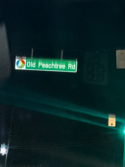 A green street sign that reads "Old Peachtree Rd" vertically is attached to a dark post. Below the text is a multicolored circular logo. Above the sign and to the left is a square grid of dark circles. To the right of the street sign are two patches of light green on the dark background.
