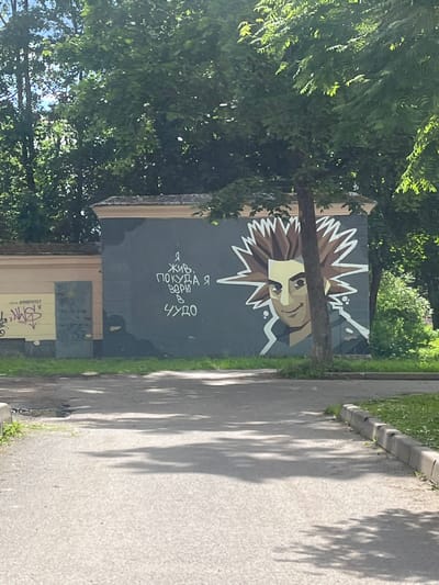 The image shows a mural on a wall in Boksitogorsk, Russia. The mural depicts a person with spiky brown hair and a yellow face, looking to the left. The person is smiling, with their eyes closed. There is text written in Russian on the wall next to the mural, which translates to "I live as long as I believe in a miracle." The wall is gray and appears to be part of a building. There are trees and foliage in the background, suggesting an outdoor urban environment. The ground in front of the wall is a paved surface, possibly a sidewalk or street, with some grass and a curb visible. The overall scene is one of urban art in a public space.
