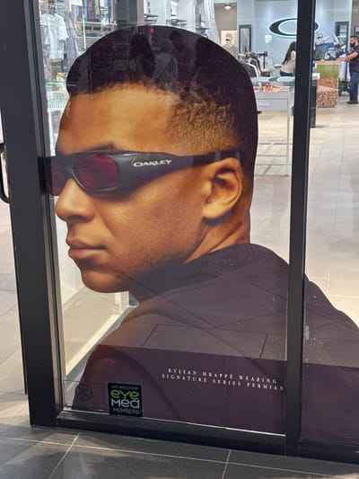 A large poster featuring French professional footballer Kylian Mbappé is displayed behind a reflective glass surface, likely at the entrance of a retail store in Schaumburg, United States of America. Mbappé is shown in a left profile, from the upper chest, wearing black Oakley sunglasses with prominent red-tinted lenses. The "OAKLEY" brand logo is visible in green on the right temple of the sunglasses. His hair is short and dark, and his skin tone is medium-brown.
Text printed in white on the lower right portion of the poster reads: "KYLIAN MBAPPÉ WEARING SIGNATURE SERIES PERMIAN". A small, square sign adhered to the lower left corner of the glass features a green logo and the text "WE WELCOME eyeMed MEMBERS".
The glass surface exhibits multiple reflections from the interior of the store and its surroundings. These reflections include overhead square light fixtures, sections of light and dark grey tiled flooring, and various retail displays. Visible through the reflections and the display are several objects: an illuminated Oakley "O" logo further inside the store, mannequins dressed in clothing, shelves holding merchandise, and a white display table with colorful items. Two individuals are also partially reflected in the background: a woman with long dark hair wearing a light-colored top, and a man with a beard wearing a dark shirt and jeans, standing near a display counter. The glass itself shows minor smudges and hairline cracks. The display is framed by a black border.
