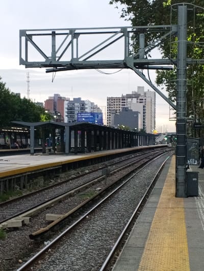 An outdoor, eye-level photograph depicts a multi-track railway station in Ramos Mejía, Argentina, under an overcast sky. In the immediate foreground on the right, a concrete train platform features a textured yellow safety strip along its edge. A dark grey metal support pole rises from this platform, affixed with a grey waste bin (marked "AKIS") and a sign displaying "Andén" (Platform). Graffiti is present on the lower section of the pole.
Multiple railway tracks, laid on ballast, extend from the lower left towards the mid-right, curving gently in the distance. Across these tracks, to the left, lies another platform with a long, dark-colored covered waiting area supported by pillars. Several individuals are seated on benches under this covered structure.
Above the tracks, a prominent, light grey metal truss structure spans the width of the railway, supported by the aforementioned pole on the right and another pole outside the frame on the left. Electrical wires hang from this overhead structure.
In the background, a dense urban landscape is visible, characterized by numerous multi-story buildings of various designs and heights. A large blue billboard is partially visible on the left, displaying "EL PRIMERO EN ARGENTINA" (The first in Argentina). Green tree foliage is present on both sides of the railway. In the far distance, an elevated roadway or bridge with visible vehicle lights can be discerned, along with green railway signal lights.