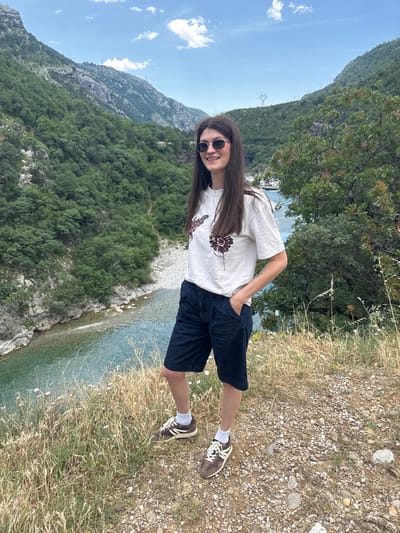 A full shot captures a young woman, likely in her early twenties, standing on a rocky terrain overlooking a river in an unspecified region of Montenegro. She has long, dark brown hair and is wearing sunglasses. She is dressed in a white t-shirt with a floral design and navy shorts, complemented by brown sneakers and white socks. Her right hand is in her pocket, and her left hand rests on her hip. The terrain beneath her feet is dry and rocky with sparse vegetation. Behind her, a turquoise river snakes through a lush, green valley, flanked by tall mountains. The sky is a pale blue with scattered clouds.