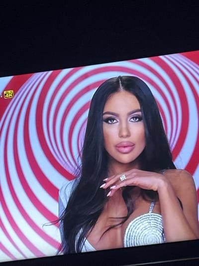 A woman with dark hair is featured against a red and white swirling background. She wears a silver, textured top and a ring is visible on her hand. The image appears to be taken of a screen displaying 4K quality. There is no evidence to support this photo was taken in Zetiovo, Bulgaria.