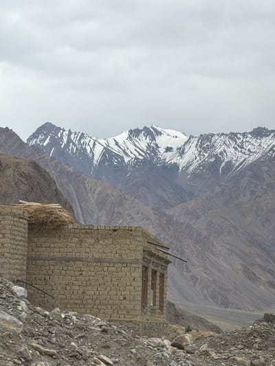 An overcast, cloudy sky hangs over a rugged landscape in Zangla, India. In the foreground, a partially constructed building made of stone bricks stands incomplete. A pile of straw and wooden beams is situated on top of one section of the wall. The building's windows are visible, some with wooden frames. A black pipe extends from the building into the surrounding terrain. The ground leading up to the structure is littered with rocks and debris. In the mid-ground, steep, barren mountains rise, their slopes textured with striations and erosion. In the background, a majestic mountain range is visible, its peaks capped with snow, creating a stark contrast with the arid lower slopes. The overall scene depicts a remote, mountainous environment with signs of human habitation in an undeveloped state.