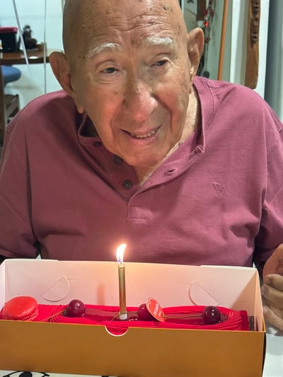 An elderly bald man with prominent white eyebrows is depicted in a medium close-up, smiling directly towards the viewer. He is wearing a maroon-colored, short-sleeved collared shirt with a three-button placket and a small embroidered logo on the left chest. In front of him, on what appears to be a table, is a rectangular dessert contained within a light brown and white cardboard box. The dessert itself is red, possibly a log or bar cake, adorned with a single lit golden candle in its center. Decorations on the dessert include a red macaron on the left, and two dark red cherries along with a small red tag on the right. The man's left hand is partially visible on the right side of the frame, resting near the dessert box. The background is blurred, showing an indoor environment with indistinct objects on shelves or furniture. The scene is located in Bnei Dror, Israel.