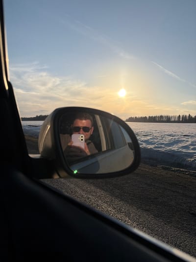 A car's side mirror reflects a person wearing sunglasses holding a purple cell phone with a double-lens camera. Outside the car window, a wintery landscape is visible with a snow-covered area and trees, under a sky with the sun visible. The location is potentially near Bol'shaya Sosnova, Russia, based on the presence of Russian text in the image's name. The car is on a paved road.