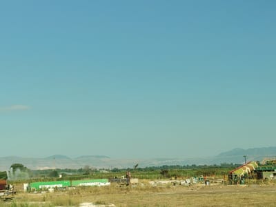 This image depicts a rural landscape under a clear blue sky. In the foreground, dry, yellowish-brown grass covers the ground. Behind this, a line of green shipping containers is visible, some bearing the word "EVERGREEN" in white lettering. Several industrial structures, including what appears to be a large water tower or tank with camouflage-like patterns, are scattered among the containers and surrounding vegetation. In the middle ground, a field of green crops, possibly corn, stretches towards distant hills. A water sprinkler is spraying water into the air in the left-center of the image, creating a misty effect. The background features a range of hazy, layered mountains. The scene appears to be located in a semi-industrial or agricultural area, likely on the outskirts of a city. There are no people visible in the image.