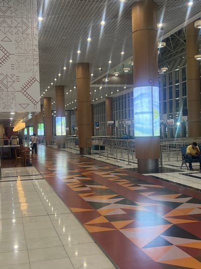 The image shows the interior of Bole International Airport in Addis Ababa, Ethiopia. The scene is viewed from a high angle, looking down a wide corridor. The corridor is lined with cylindrical columns covered in a light brown material, some of which have digital screens displaying images. The floor has a geometric pattern in various shades of brown, orange, white, and black. The ceilings are high with a grid-like structure and evenly spaced recessed lights. There are people seated in chairs along the side of the corridor. A man is visible in the upper part of the image, sitting in a waiting area. The floor is tiled with a light-colored material that reflects light. In the bottom left corner, there is a section of wall with geometric patterns in white and brown.