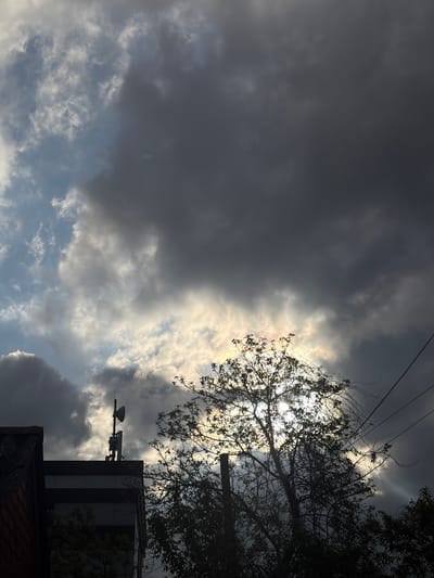The image depicts a cloudy sky above a building with trees silhouetted against the light. The sky is filled with dark, heavy clouds on the left and lighter clouds with patches of blue sky on the right. In the upper right, tree branches partially obscure a bright light source, likely the sun. A building is visible in the lower right, with a weather instrument mounted on its side. Powerlines run along the top of the image. The location is unknown and unconfirmed.