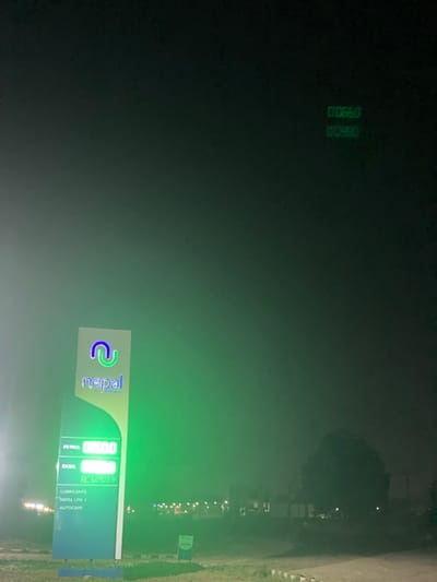 The image presents a nighttime view of a "Nepal" fuel station located in Issele Uku, Nigeria. A tall, illuminated pylon sign, positioned slightly to the left of the frame's center, displays the "Nepal" brand with a blue and green looped logo. Below the logo, digital readouts indicate fuel prices: "PETROL 0950.0" and "DIESEL 0980.0". Additional text on the sign lists "LUBRICANTS", "NEPAL LPG / AUTOCARE", and "RC 60937". The ground directly in front of the sign appears to be a paved or dirt surface. In the background, a line of distant artificial lights is visible, suggesting other structures. A large, dark tree silhouette occupies the right side of the frame. The dark sky above is permeated with a green ambient glow emanating from the bright sign. Faintly discernible in the upper right portion of the sky are two sets of green digital characters, "00660" and "00980". No people or vehicles are visible in the scene.