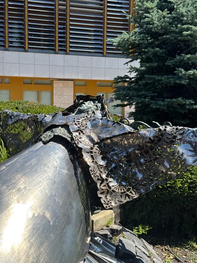 The image shows a sculpture against the backdrop of a building and trees. The sculpture is made of polished metal, possibly stainless steel, and has a lace-like, decorative overlay. The building in the background is multi-story, with a facade of alternating panels of light-colored stone or tile and sections with multiple narrow windows or louvers. A dense tree with bluish-green foliage stands to the left of the building. The sculpture appears to be located outdoors in a landscaped area. Without additional information, a precise location within Siófok, Hungary cannot be determined.