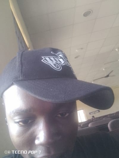 A medium shot photograph features a person wearing a black baseball cap with a white logo. The cap covers most of the person's head, obscuring their hair and forehead. Only the person's eyes, nose, and part of their cheek are visible. The background suggests an indoor setting with a tiled ceiling, lighting fixtures, and ceiling fans. In the distance, rows of empty seats are visible, characteristic of an auditorium or lecture hall. A window with natural light and a sign with text are also discernible in the background. The bottom of the image contains a watermark that reads "TECNO POP 7". The location of Eleweran, Nigeria, is not visually verifiable from the image content.