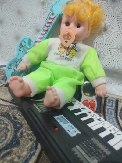 A doll with blond hair, a pacifier, and a light green outfit sits on a toy keyboard. The doll is positioned with its legs extended and feet resting on the keyboard. To the left of the doll, a blue toy guitar with pink buttons is visible. Behind the doll and guitar is a patterned wallpaper. A rug with a blue and brown pattern lies beneath the keyboard and doll. The scene appears to be indoors, possibly in a child's room or play area, in the city of Cholpon, Kyrgyzstan.