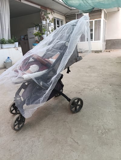 A stroller with a baby inside is covered by a white mosquito net, placed on a concrete surface outside a building. The stroller is dark gray with black wheels. The baby appears to be sleeping, covered by a maroon blanket and white clothing. The baby's legs are visible, and one leg seems to have a bandage.
Behind the stroller, there's a white door with glass panels and a white frame, leading into a building. To the left of the door, a white curtain hangs. Further left, there are several potted plants on shelves and a white countertop. A large blue water jug is visible on the left side of the image.
The ground is made of rough concrete. Above the scene, a green mesh-like fabric is stretched, likely providing shade. The overall setting appears to be outdoors, possibly a courtyard or patio area. The lighting suggests it is daytime. The presence of the mosquito net indicates a climate where insects are a concern. The environment seems to be residential. No text is visible in the image.