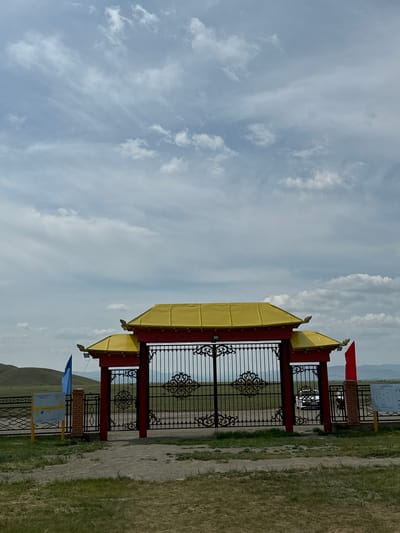 The image shows an ornate gate with red pillars and a golden, pagoda-style roof. The gate is made of black wrought iron with intricate scrollwork. On either side of the gate, there are brick pillars. A blue flag flies on the left side, and a red flag flies on the right side. Behind the gate, a white car is visible. The landscape behind the gate is open and hilly, with distant mountains under a cloudy sky. The foreground is a grassy area with a dirt path. Informational signs are posted on either side of the gate. The location is the entrance to the Orongoi Buddhist Temple complex in Orongoi, Russia.