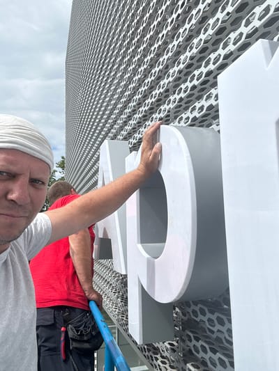 The image shows two people working on a large, white, three-dimensional number sign. One person, wearing a red t-shirt and dark work pants, is holding a blue pole and appears to be in the process of installing or adjusting the sign. The other person, wearing a grey t-shirt and a white head covering, is partially visible in the foreground, looking towards the camera with a neutral expression. The background features a textured, grey and white geometric pattern, possibly part of a building facade. The lighting suggests it is daytime. The scene appears to be outdoors, with a hint of trees and a cloudy sky in the background. The image is oriented vertically. There is no visible text in the image.