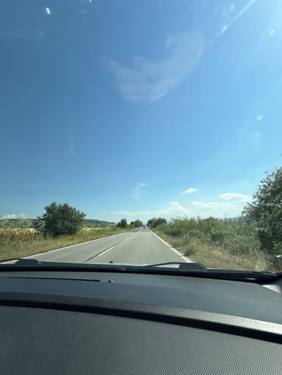 The image captures a first-person perspective from inside a vehicle traveling on an asphalt road in Mursalevo, Bulgaria, under a clear, bright blue sky with scattered white clouds. The road, featuring a solid white line on the right shoulder and a broken white center line, extends into the distance, where a light-colored vehicle is faintly visible. To the left, fields of golden crops are interspersed with green trees and bushes, with distant hills under the horizon. The right side of the road is bordered by dense, green wild vegetation and scattered trees. The lower portion of the image shows the dark gray dashboard of the vehicle, which has a textured pattern, and the base of the windshield wipers. Reflections and minor imperfections are visible on the windshield in the upper sky area. No individuals are explicitly present within the frame.