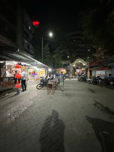 This night-time image depicts a commercial street scene in Surat, India. The foreground is paved with grey interlocking blocks, displaying the distinct shadow of a person, likely the photographer, extending from the bottom center. On the left, a row of brightly lit street stalls operates under awnings. At one stall, two men are visible: a vendor behind the counter and a customer holding several bright orange/red hanging items. Gujarati script, including "શ્રી ક્ષેત્રપાલ," is visible on the shop signage. Numerous motorcycles and scooters are parked along both sides of the street. In the middle ground, a man in a light-colored plaid shirt stands with his hands on his hips, positioned near a small metal table and several parked motorcycles. Further down the street, a decorative stone archway, indicative of a temple entrance, is prominently visible. Additional stalls and vehicles are present beyond this archway. The background features multi-story buildings, one on the left exhibiting a partial red neon sign displaying "ON." Multiple streetlights illuminate the environment, creating distinct shadows. Trees are integrated into the urban landscape on the right side and in the distance. The scene suggests active night-time commerce within a temple precinct.