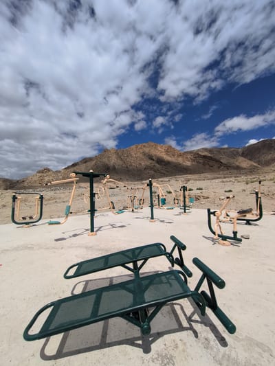 This is an outdoor scene in Phyang, India, featuring an exercise park situated in a arid, mountainous region. The foreground is dominated by two dark green, metal exercise benches with perforated seating surfaces and curved support legs. In the middle ground, a variety of outdoor fitness equipment is arranged on a light-colored paved surface. These machines, primarily in a tan and green color scheme, include leg press machines, chest press machines, and other strength-training apparatuses. The equipment is spaced apart, suggesting a designated workout area. In the background, the rugged, brown, and rocky mountains rise under a bright blue sky filled with scattered white clouds. The overall impression is one of an open-air gym set against a stark, natural landscape. There are no people visible in the image.