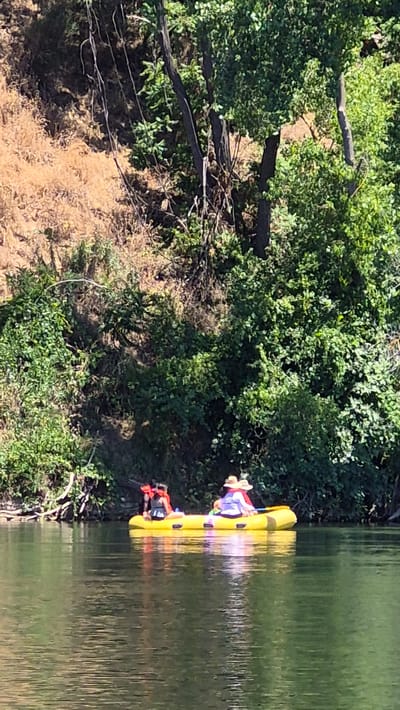 In Carmichael, California, a bright yellow raft carrying two individuals navigates a calm river on a sunny day. The raft is positioned on the water's surface, with its inhabitants facing away from the camera. One person, situated towards the front of the raft, is wearing an orange life vest over a black top. The second individual, positioned behind the first, wears a wide-brimmed straw hat and a light-colored top with red accents, and holds a paddle. The riverbank in the background is densely covered with lush green foliage and trees, interspersed with dry, brown brush on a steep incline. The water reflects the surrounding greenery and the sky.