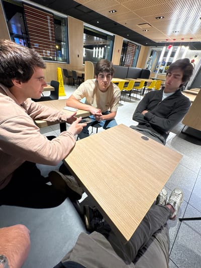 The scene is an interior view of a modern McDonald's restaurant in South Bend, United States, likely during evening or nighttime hours, indicated by artificial lighting and exterior reflections through windows. The establishment features light wood-grain laminate tables, black seating booths, and individual chairs with black frames and some yellow seats. The ceiling is light-colored and perforated, equipped with recessed lighting. The floor is covered with light grey tiles. A yellow caution cone is visible in the background.
Four individuals are present. Three males are fully visible, seated at two separate light wood-grain tables. The fourth individual is partially visible in the immediate foreground, indicating the perspective of the photographer.
- **Subject 1 (Left):** A male with brown hair, wearing a light brown hooded sweatshirt. He is seated at a square table, holding a dark smartphone horizontally in his left hand and gesturing with his right hand while looking towards Subject 2.
- **Subject 2 (Center):** A male with brown hair, wearing a light beige t-shirt and blue jeans. He is seated at the same table as Subject 1, facing him, with his hands clasped together on the table. He is looking towards Subject 1. A light-colored smartphone rests on the table in front of him.
- **Subject 3 (Right):** A male with brown hair, wearing a dark grey zippered long-sleeved top over a white collar. He is seated at a separate square table, his arms crossed, and his head tilted with eyes appearing closed or downcast, indicative of rest or fatigue.
- **Subject 4 (Foreground):** Only the lower legs, clad in dark trousers and light-colored sneakers, and a left forearm with a metallic watch are visible in the lower left foreground.
Subjects 1 and 2 appear engaged in a verbal exchange. Subject 3's posture suggests disengagement from the immediate interaction.