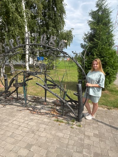 A young woman with blonde hair stands to the right of a metal art installation and a large evergreen tree. She is wearing a light green t-shirt and shorts, and white clogs. The metal art piece is in the foreground, featuring a rustic, tree-like structure with various metal elements, including what appears to be letters spelling out "BREYTOBO" arching over the top. The background shows green trees, a cloudy sky, and some distant power lines. The scene is set on a paved walkway made of rectangular stones.