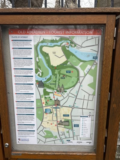The image shows a wooden-framed tourist information board displaying a map of Old Aberdeen, a historical part of Aberdeen City, United Kingdom. The map outlines key points of interest, including the Brig o' Balgownie, Seaton Park, St Machar's Cathedral, the Chanonry, Benholm's Lodging, Cruickshank Botanic Garden, the Zoology Museum, the Old Aberdeen Town House & Museum, the Duncan Rice Library, King's College & Chapel, the Powis Gateway, and Crombie Halls of Residence. The map also indicates the locations of various shops and services such as cafes, takeaways, pubs, churches, mosques, bookshops, petrol stations, general stores, cash machines, and chemists. The information board features text descriptions for each point of interest, providing historical context and details. The River Don winds through the map, and the University of Aberdeen logo is displayed at the bottom. The environment around the board includes a gray metallic object.