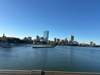 This image depicts a wide panoramic view of the Boston skyline across the Charles River, observed from an elevated position in Cambridge, Massachusetts, likely from the Longfellow Bridge. A dull grey metal railing occupies the lower portion of the foreground. The Charles River fills the midground with calm, rippled, deep blue-green water. On the left bank, numerous white sailboats are moored in a marina, backed by buildings with dark roofs and dense green foliage. A prominent dark steeple rises above the trees on the far left. A small, tree-covered island is visible in the river slightly to the right of the image center. The background showcases the Boston skyline under a clear, cloudless light blue sky. Key structures include the reflective blue-green John Hancock Tower (200 Clarendon Street) and the lighter-colored Prudential Tower, along with numerous other high-rise and mid-rise buildings. The distinct grey dome of the MIT Great Dome is partially visible on the left side of the Boston bank amidst trees and other structures. No people or discernible actions are visible in the scene.
