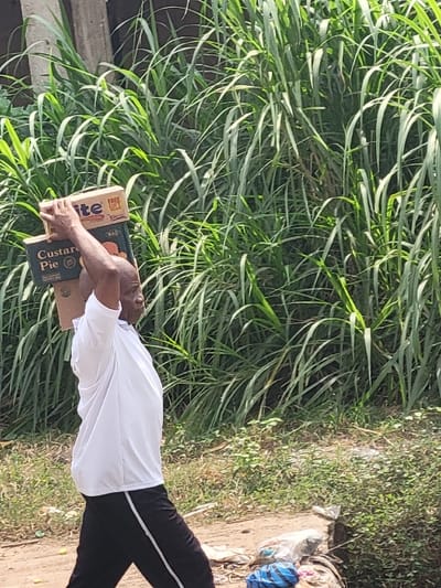 An adult male with dark skin and a shaved head is depicted walking outdoors in Puerto López, Colombia, from right to left across the frame. He wears a white short-sleeved athletic shirt and dark pants with a white stripe down each side. He is carrying two cardboard boxes stacked vertically on his head, steadying them with both hands raised, palms facing upwards. The top box is light brown and shows partially legible text including "lite" and "FREE GIFT." The bottom box is darker, featuring text that reads "Custard Pie" and a circular logo. A tattoo is visible on his left forearm. The background consists of dense, tall green vegetation, resembling reeds or sugarcane, covering the upper and right portions of the image. The ground is an unpaved dirt path with sparse green plants. To the right of the man, a pile of assorted refuse, including plastic bags, is visible on the ground. A vertical wooden post is partially visible behind the tall vegetation on the upper left. The scene is brightly lit by natural sunlight.