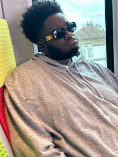 A male individual is depicted seated within a moving conveyance, likely a train, in Colombes, France. He is positioned in profile, facing slightly right, with his head tilted upward. The subject has dark skin, a short dark beard, and short, dark, curly hair. He is wearing a light gray hooded sweatshirt and dark sunglasses featuring a prominent gold-colored medallion on each temple arm. His lips are closed, suggesting a neutral or contemplative expression. To his left, the back of a seat with a bright red and yellow patterned fabric is visible. Through the window on his right, a blurred exterior environment is seen, including a light-colored residential building with a tiled roof and multiple windows, consistent with European architecture, against a light sky, indicating the vehicle is in motion.
