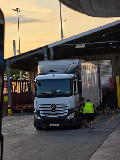 At a logistics facility in Walldorf, Germany, a white Mercedes-Benz Actros semi-trailer truck is parked next to a loading bay under a covered structure. Two men are standing near the truck; one, wearing a black t-shirt and dark shorts, is leaning against the truck's cab, and the other, wearing a neon yellow safety vest and dark shorts, is facing away from the camera and appears to be interacting with the truck. The truck's trailer is white and has the number "5070" visible on its side. The sky behind the truck shows a sunset with soft orange and yellow hues, with some scattered clouds. The ground is asphalt, marked with yellow lines. In the background, there is a large corrugated metal roller door and various industrial elements, including yellow bollards and red crates. The scene is captured during twilight.